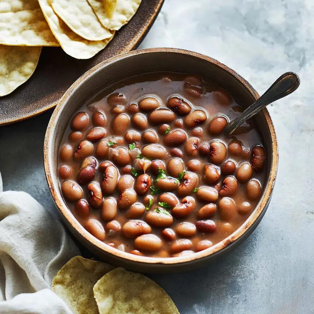 A wooden bowl filled with pinto beans in broth, garnished with chopped cilantro, alongside a plate of tortilla chips.