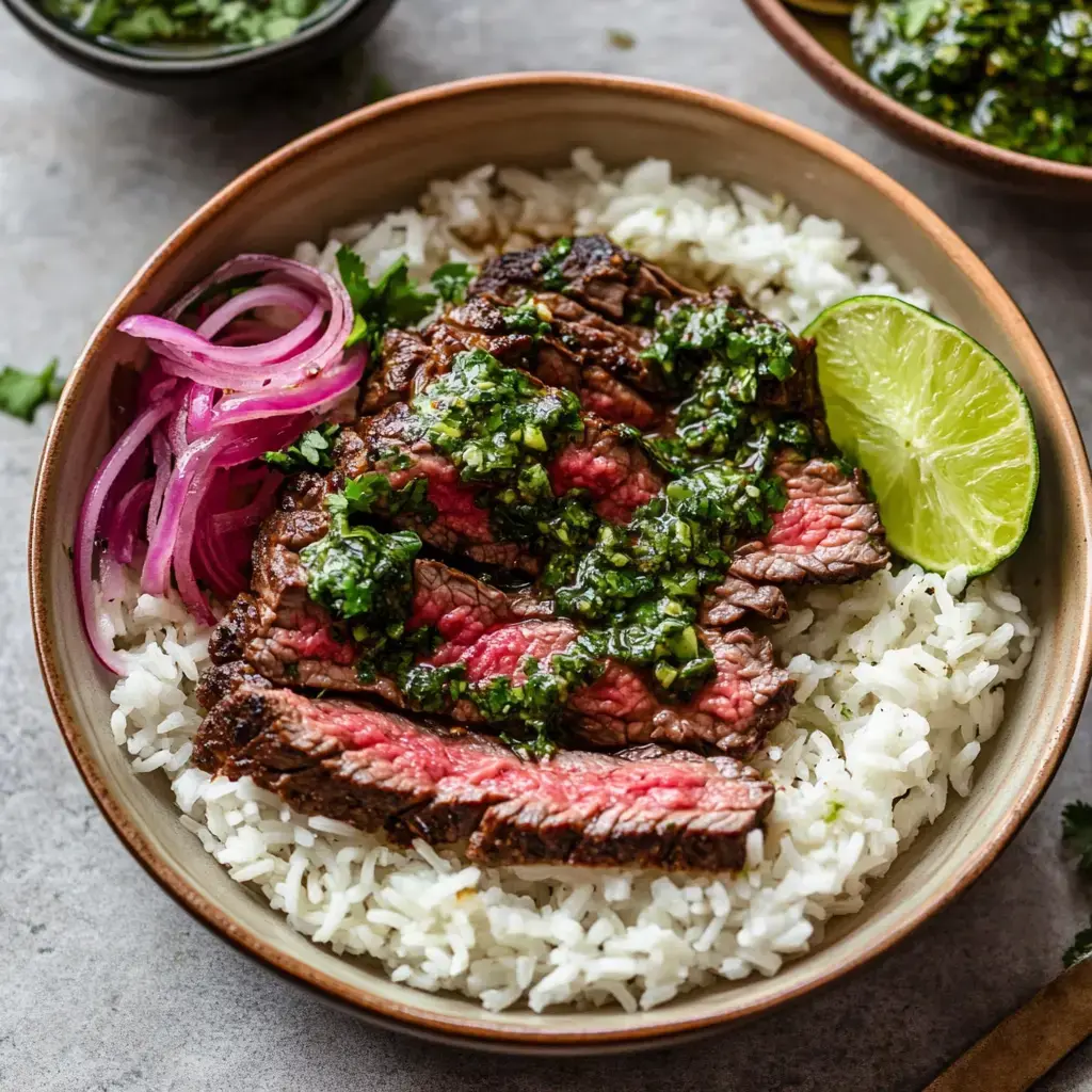 A bowl of rice topped with sliced steak, chimichurri sauce, pickled red onions, and a lime wedge.