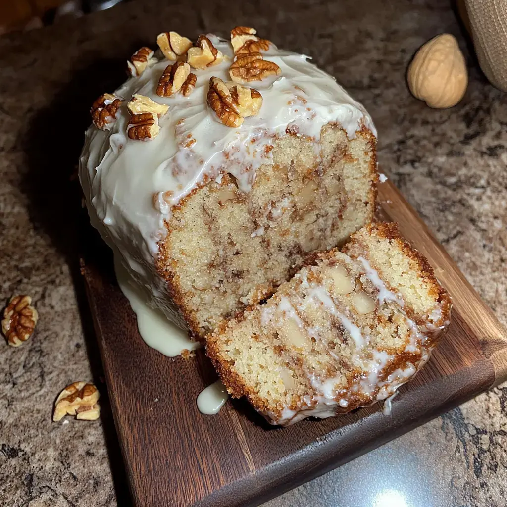 A sliced loaf cake topped with cream frosting and garnished with pecans is displayed on a wooden cutting board.