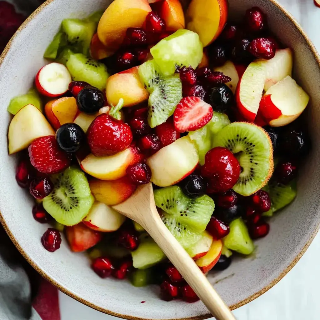 A vibrant fruit salad featuring slices of apples, strawberries, kiwi, and melons, mixed with raspberries and pomegranate seeds in a bowl with a wooden spoon.