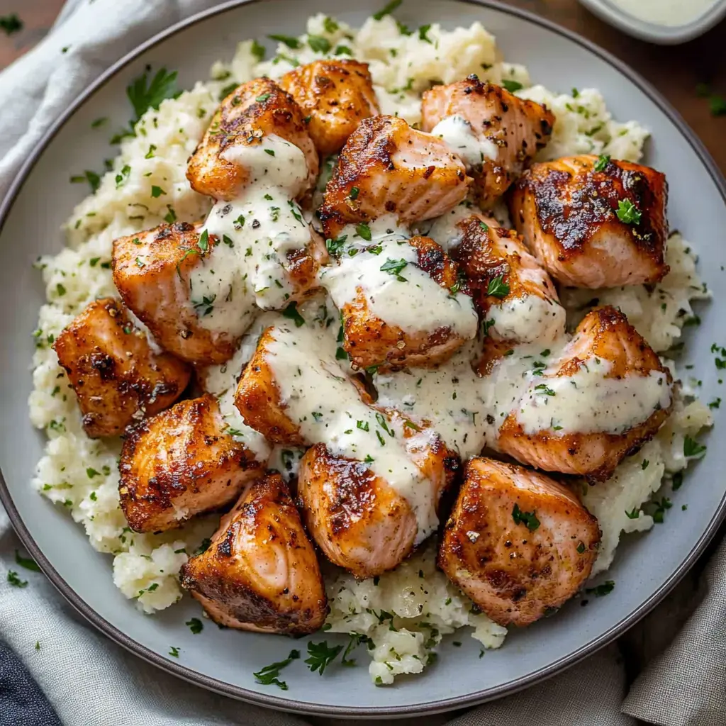 A plate of grilled salmon pieces topped with creamy sauce, served over a bed of fluffy rice and garnished with parsley.