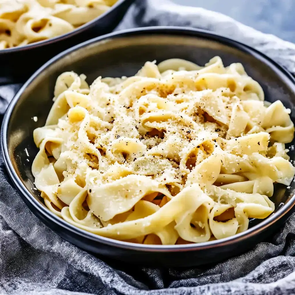 A bowl of fettuccine pasta topped with grated cheese and black pepper, served on a textured gray cloth.