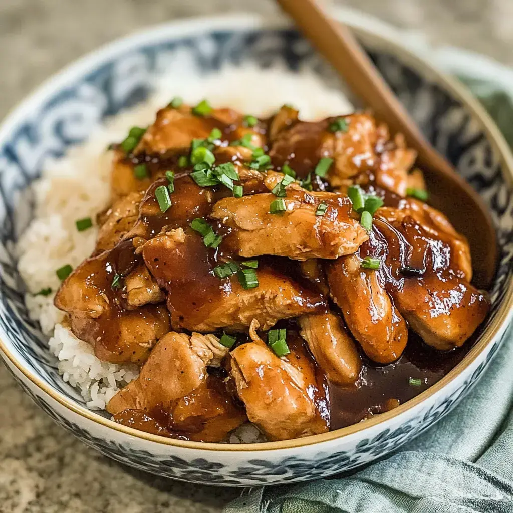 A bowl of rice topped with succulent chicken pieces covered in a glossy brown sauce and garnished with chopped green onions.