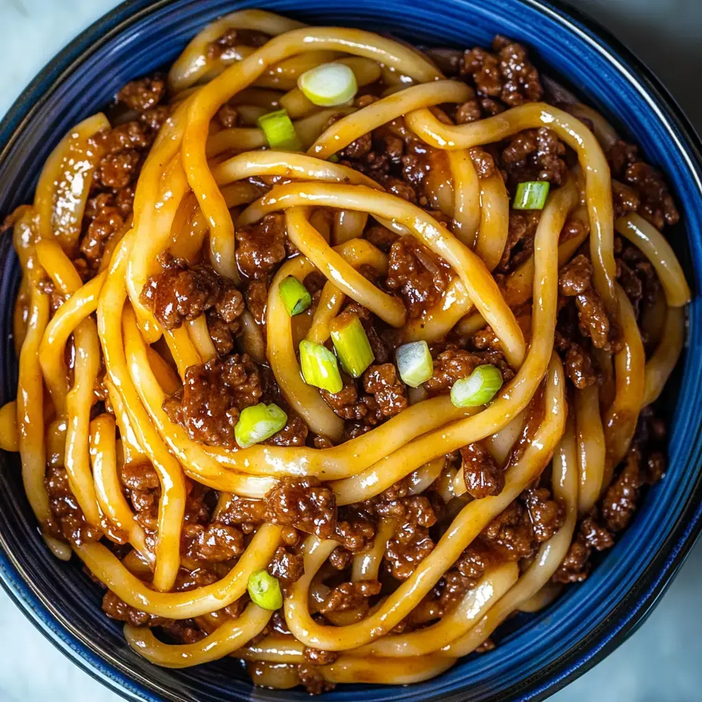 A close-up of a bowl of thick noodles mixed with ground meat, garnished with green onions.