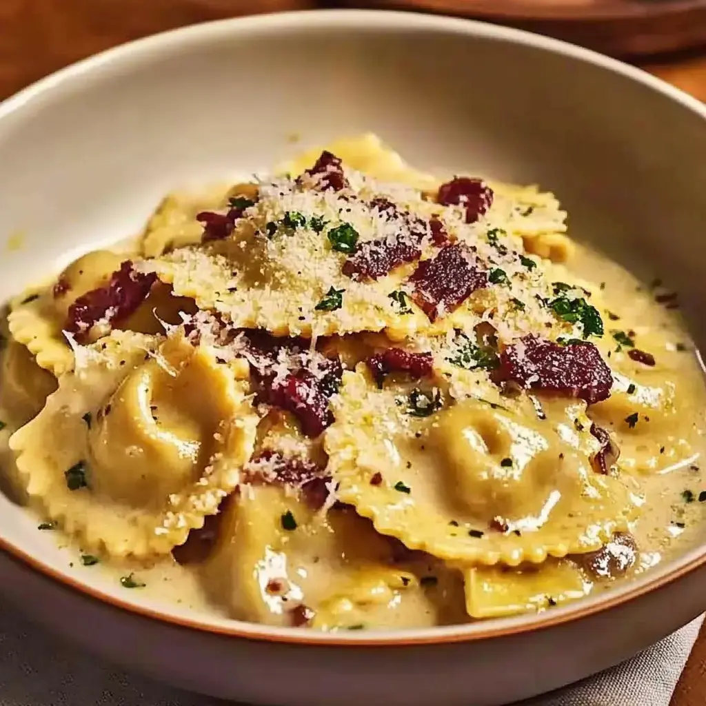 A bowl of ravioli in a creamy sauce, topped with crispy bacon bits and grated cheese, garnished with green herbs.