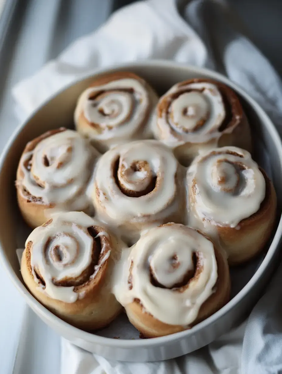 A close-up view of freshly baked cinnamon rolls topped with creamy icing, arranged in a round white dish.