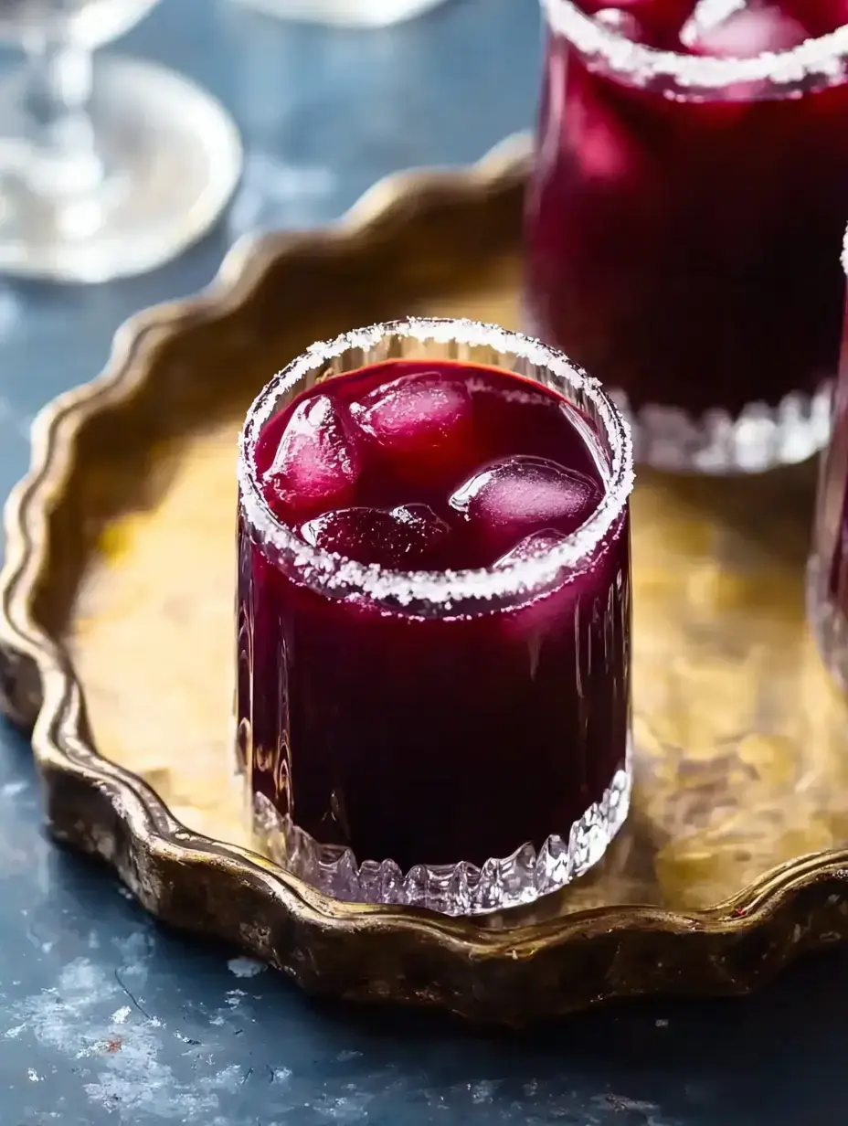 Three glasses filled with a deep purple drink and ice, garnished with salt around the rim, are arranged on a decorative gold tray.