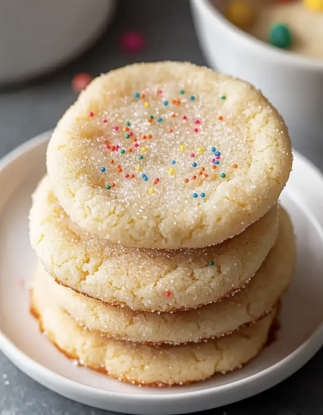 A close-up of a stack of four sugar cookies sprinkled with colorful nonpareils on a white plate.