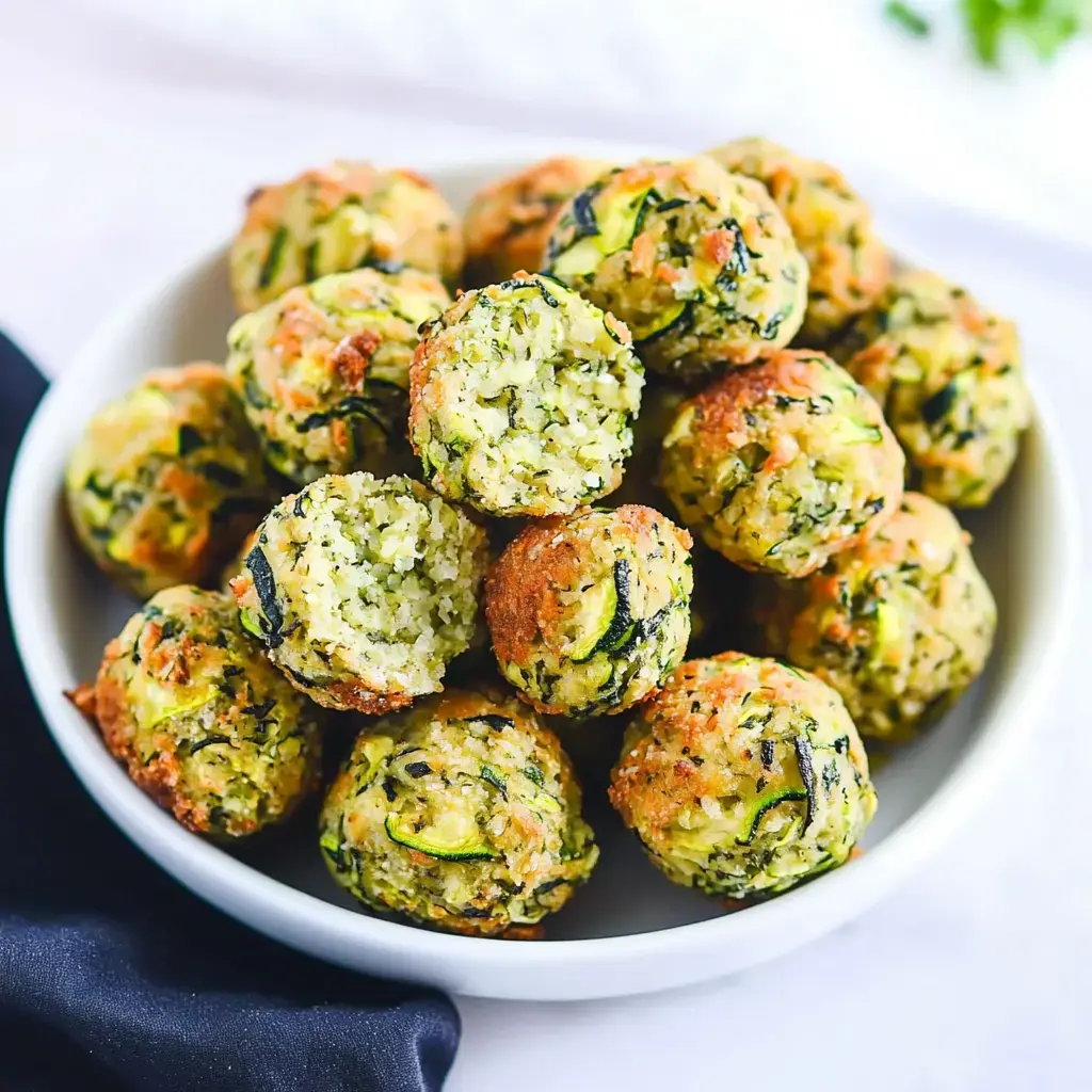 A bowl of golden-brown zucchini balls, some partially bitten, showcasing a crispy exterior and a green, veggie-filled interior.