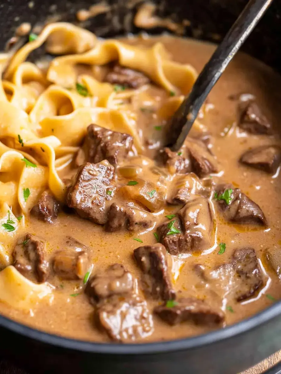 A close-up of a bowl containing tender beef chunks and wide egg noodles in a creamy sauce, garnished with parsley.