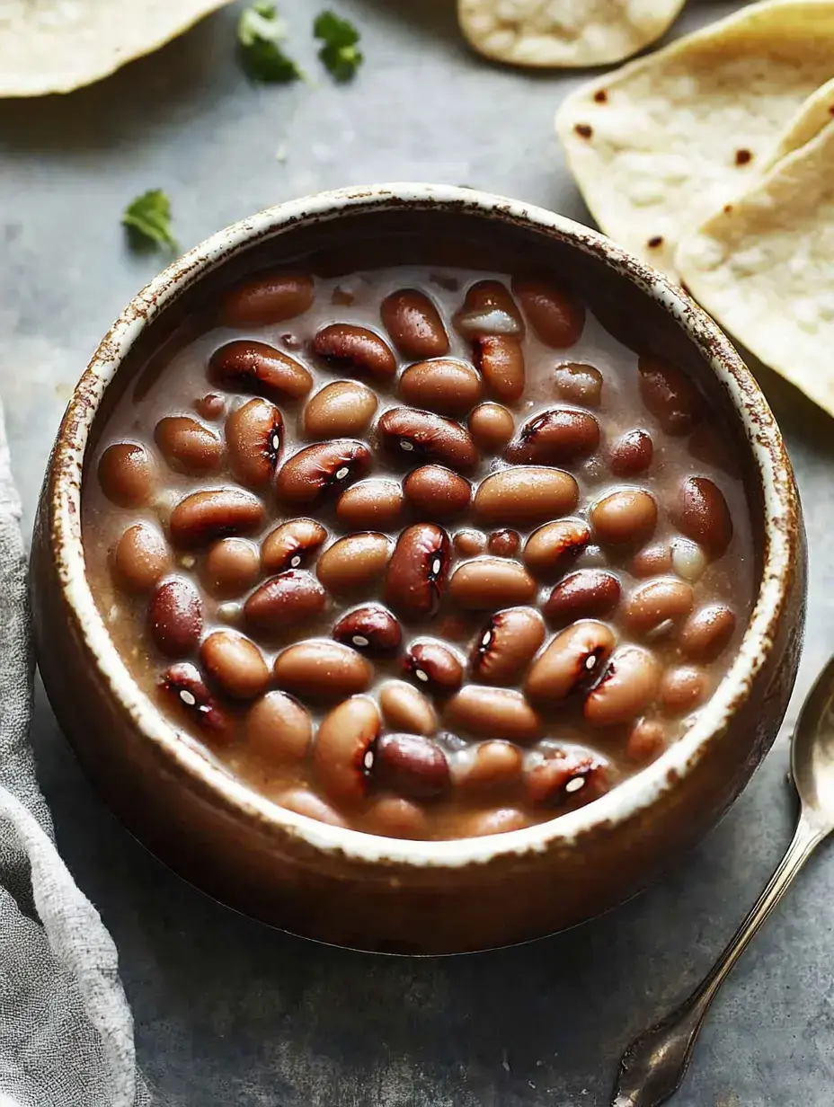 A bowl of cooked beans in broth is surrounded by tortillas and fresh cilantro on a rustic surface.