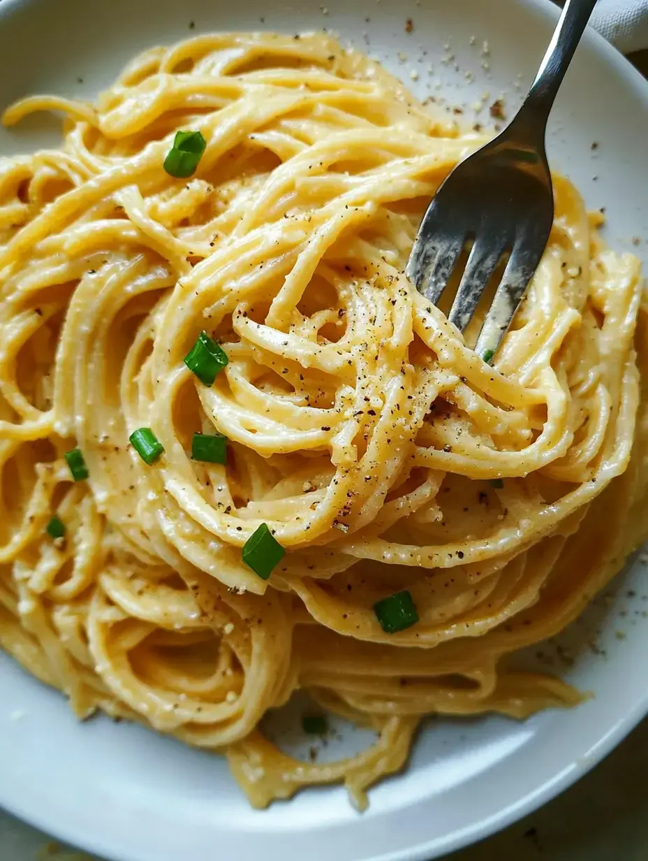 A close-up of creamy spaghetti topped with green onions and black pepper on a white plate, with a fork resting in the noodles.