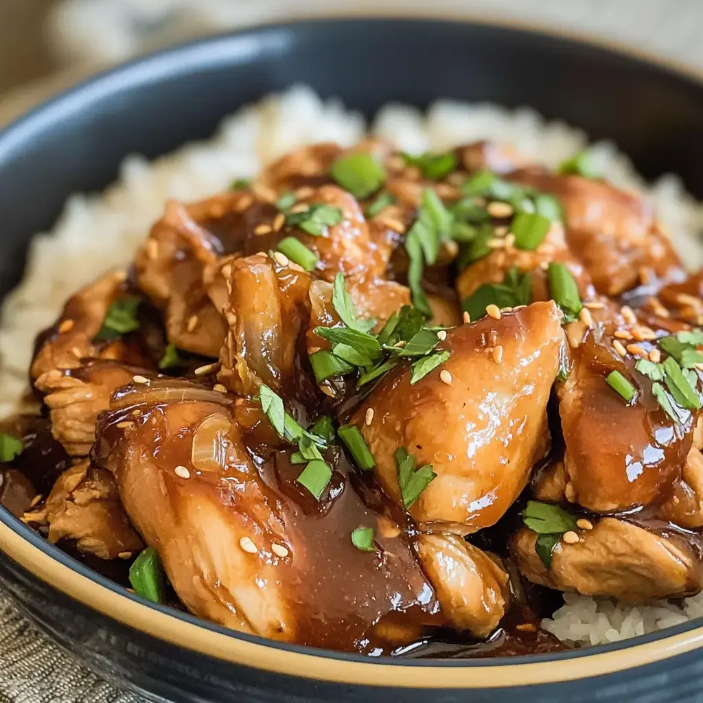 A close-up of a bowl of rice topped with tender chicken pieces in a glossy sauce, garnished with sesame seeds and chopped green herbs.