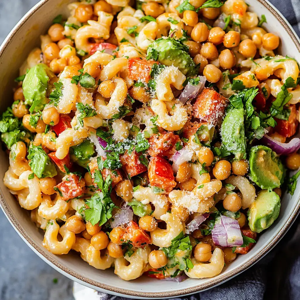 A colorful bowl of pasta salad featuring macaroni, chickpeas, diced red bell pepper, avocado, red onion, and fresh herbs, topped with grated cheese.