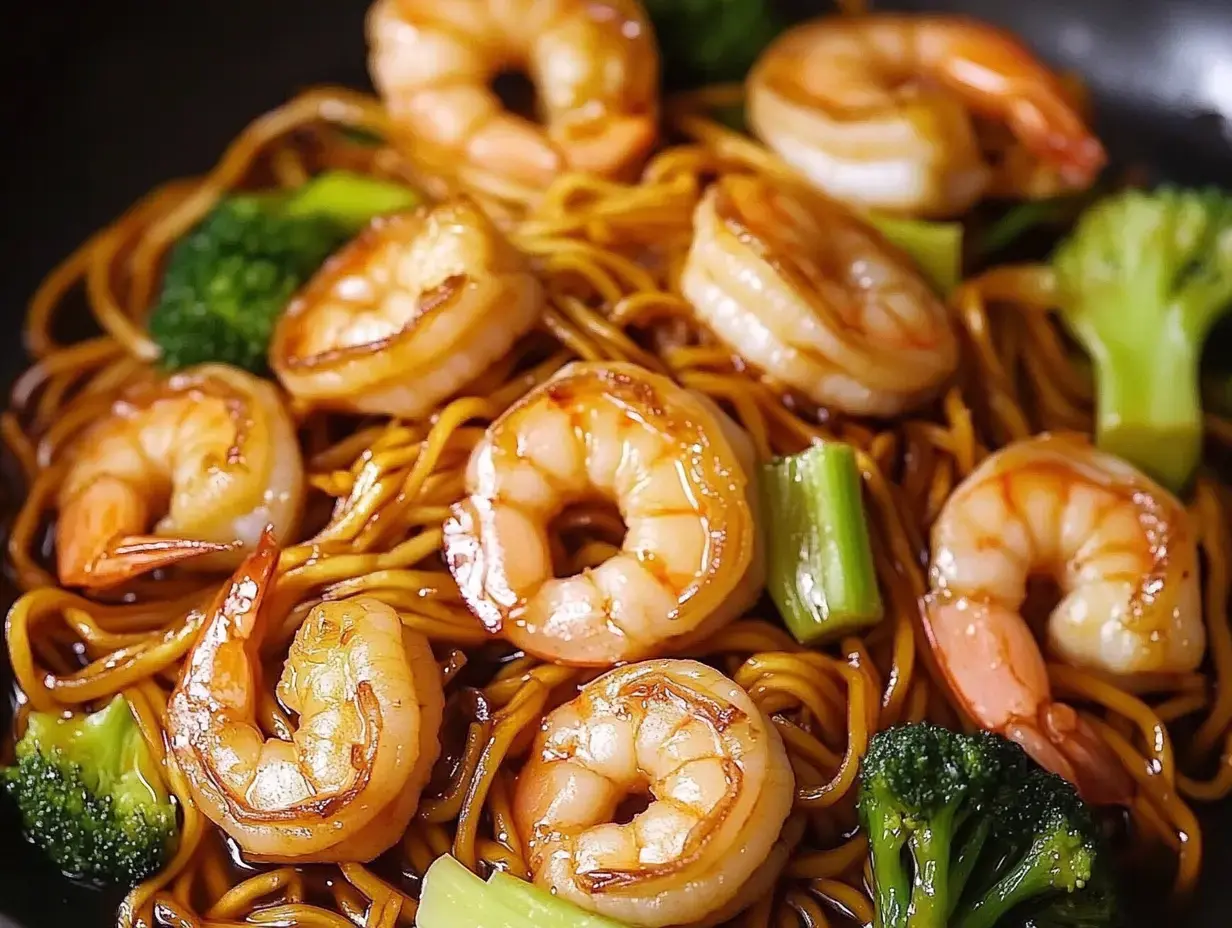 A close-up of a dish featuring stir-fried noodles topped with shrimp and garnished with broccoli.