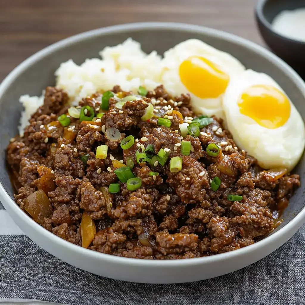 A bowl of seasoned ground meat topped with green onions and sesame seeds, served with rice and two soft-boiled eggs.