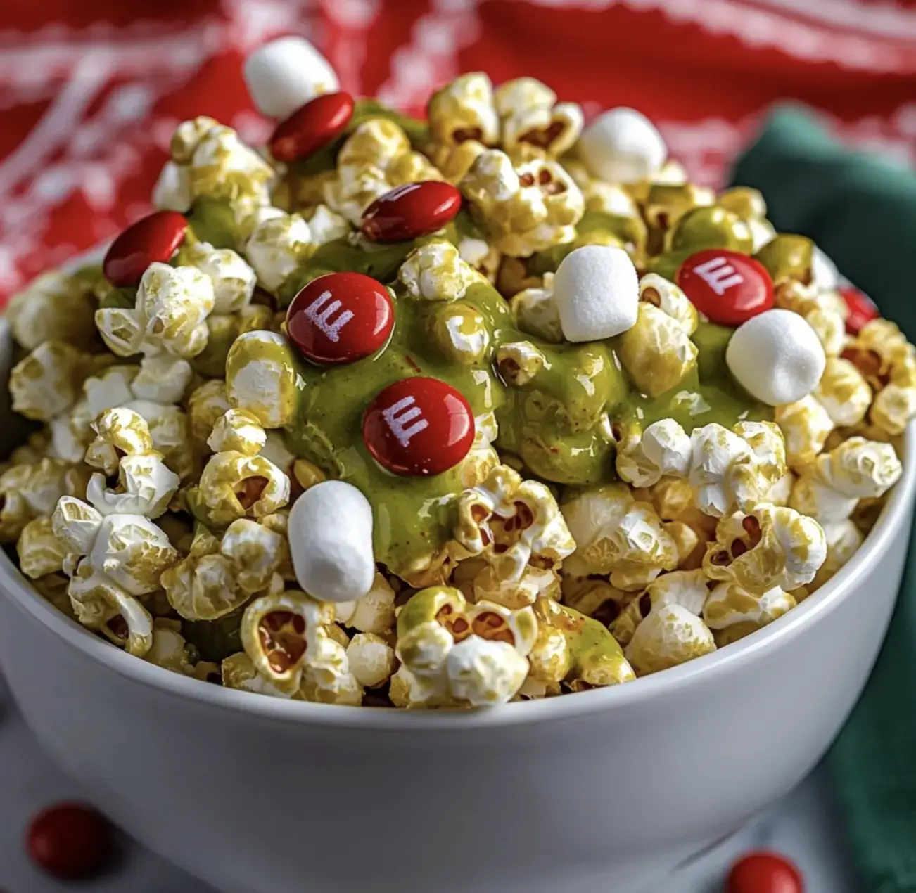 A bowl of popcorn is topped with green drizzle, red M&M's, and marshmallows, creating a festive snack mix.