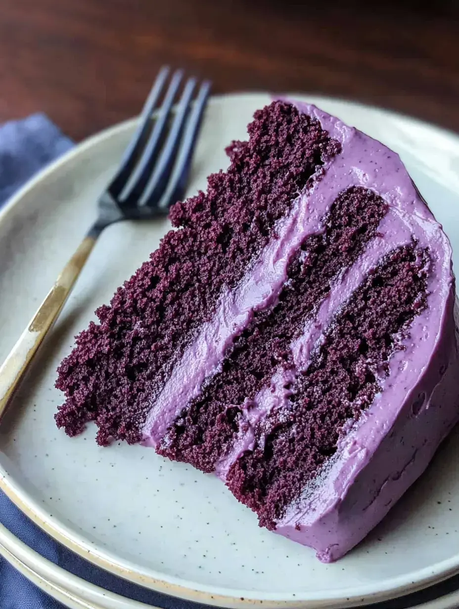 A slice of purple cake with cream frosting sits on a plate next to a fork, showcasing its moist layers.