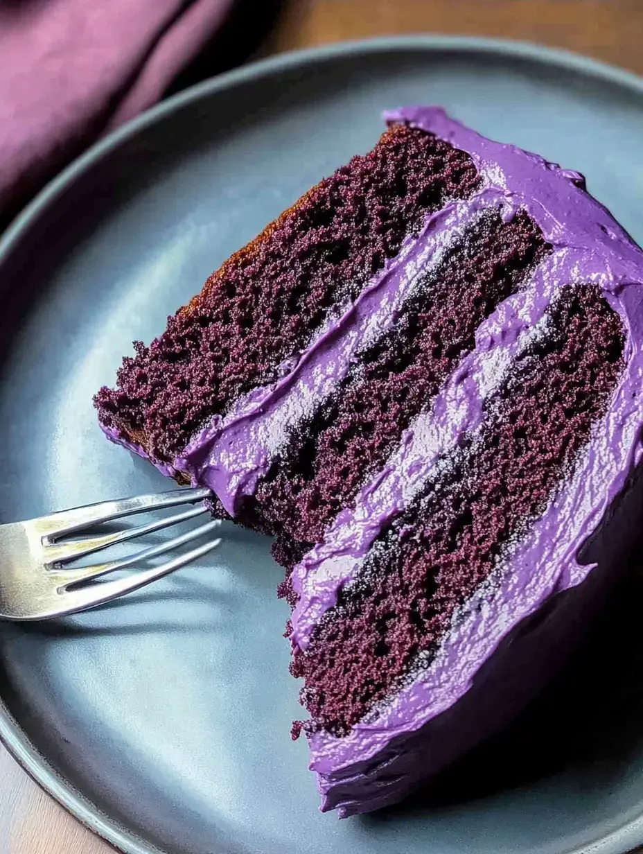 A slice of purple frosted cake is displayed on a dark plate with a fork resting beside it.