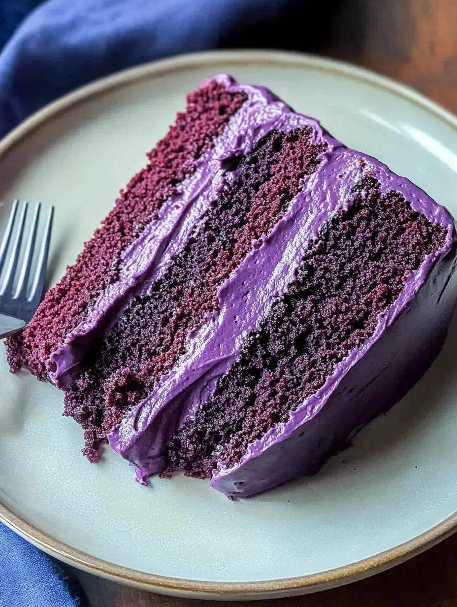 A slice of purple velvet cake with purple frosting is served on a plate alongside a fork.