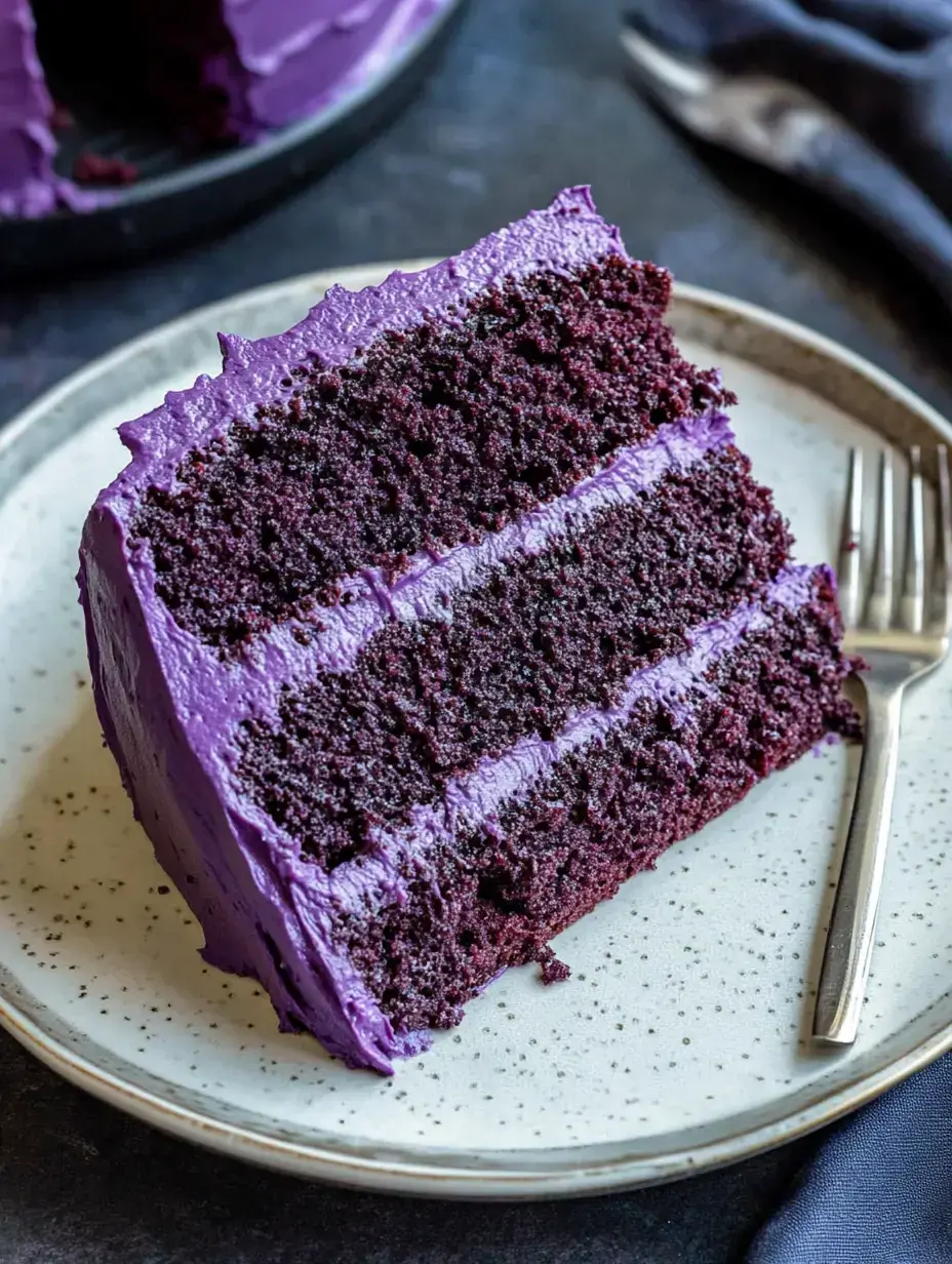 A slice of purple velvet cake with layers of dark chocolate and purple frosting on a speckled plate.