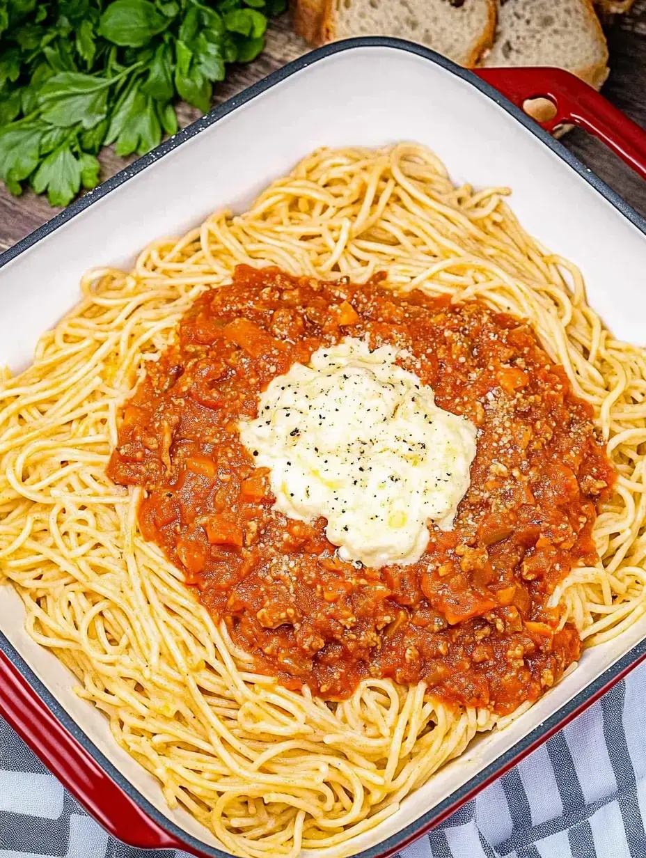 A close-up view of spaghetti topped with meat sauce and a dollop of creamy sauce, served in a square dish with fresh parsley and bread in the background.