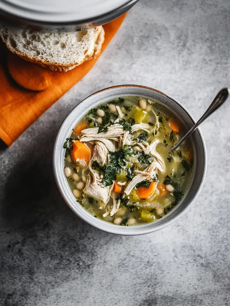 A bowl of chicken soup with shredded chicken, carrots, greens, and white beans, accompanied by a slice of bread on a textured gray surface.