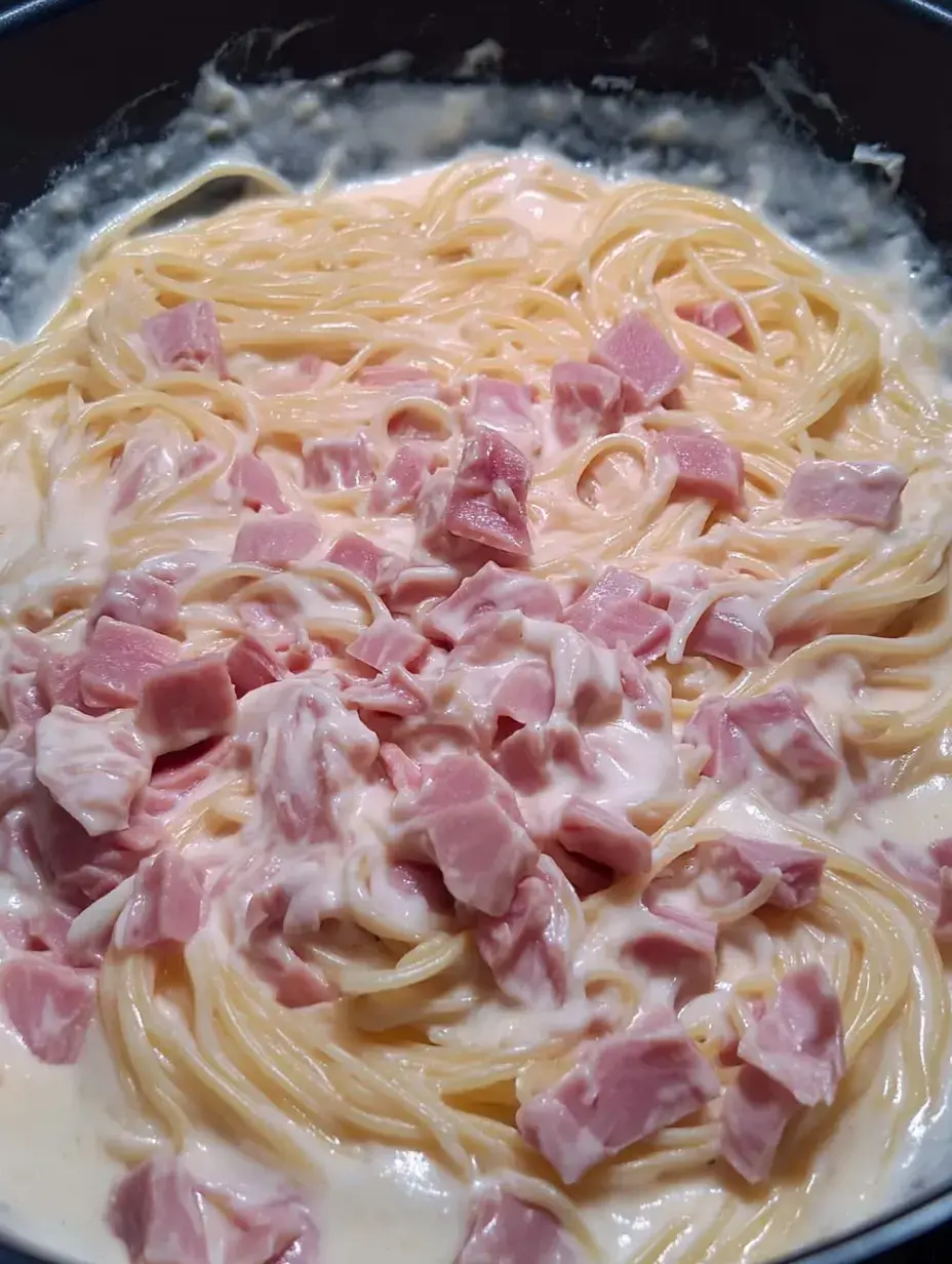 A close-up image of creamy pasta mixed with pieces of ham in a dark bowl.