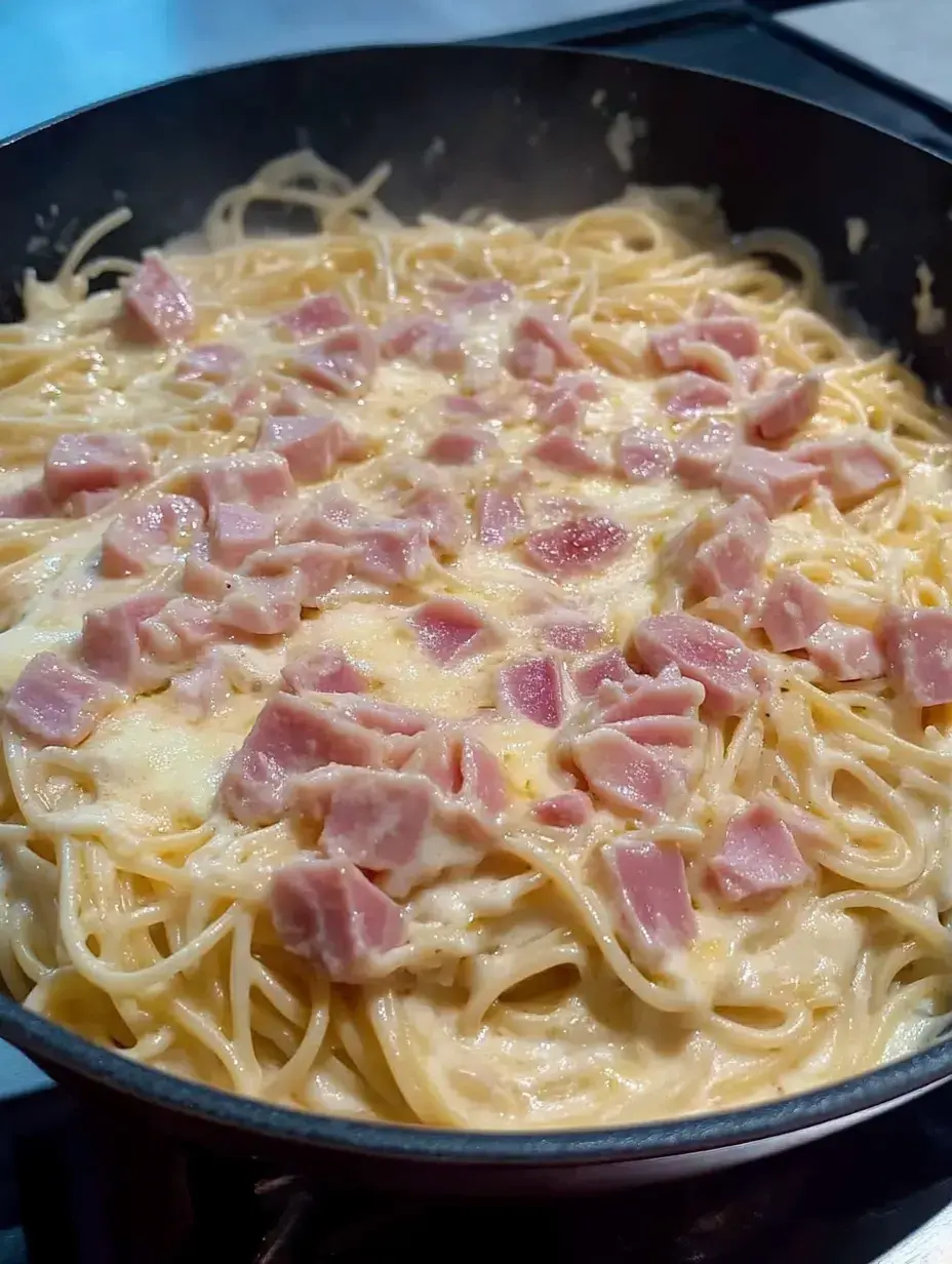 A close-up of a pan filled with spaghetti coated in a creamy sauce and topped with diced ham.