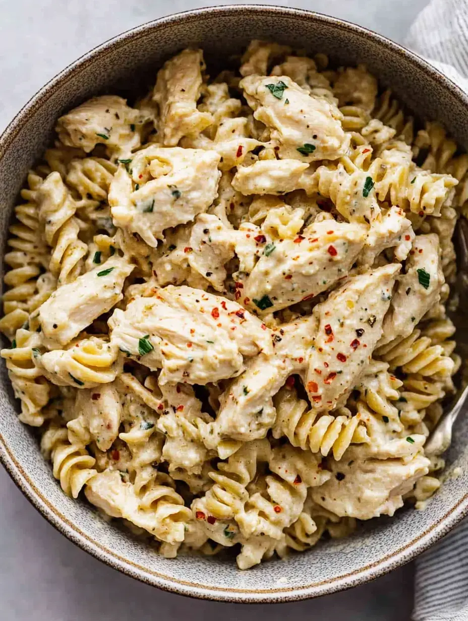 A bowl of creamy chicken pasta with rotini noodles, garnished with red pepper flakes and herbs.