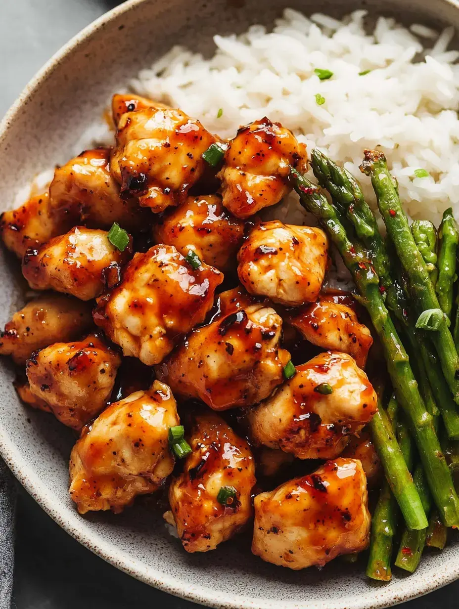 A bowl filled with glazed chicken pieces, served alongside white rice and green asparagus.