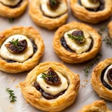 A close-up of golden, flaky pastry tarts filled with dark fruit compote, topped with a creamy layer and a dried fig slice, garnished with fresh thyme.