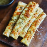 Four golden-brown cheese breadsticks sprinkled with herbs, arranged on a wooden cutting board.