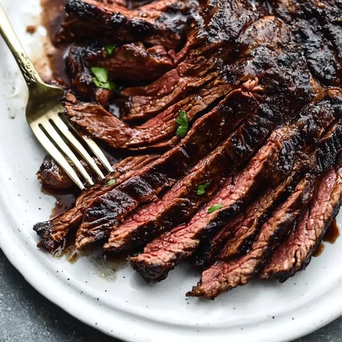 A close-up of sliced grilled beef brisket on a white platter, garnished with parsley and served with a gold fork.