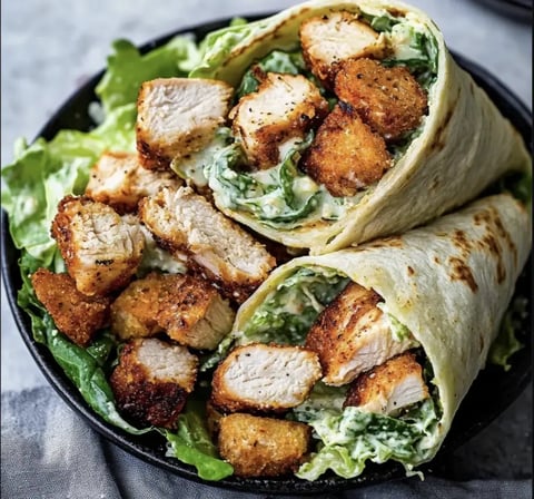 A close-up of two chicken wraps filled with lettuce and creamy sauce, served in a dark bowl on a textured surface.