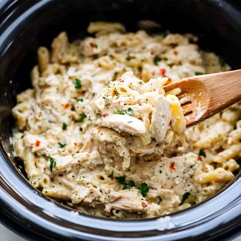 A wooden spoon is lifting creamy chicken and pasta from a slow cooker filled with a cheesy dish garnished with herbs.